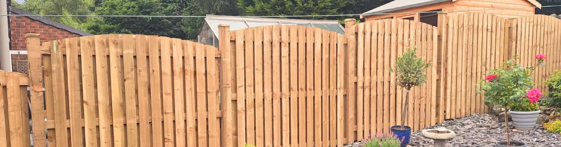 A wooden garden fence with curved tops separates a landscaped yard with gravel, potted plants, and blooming flowers from a background of trees, a shed, and a greenhouse.