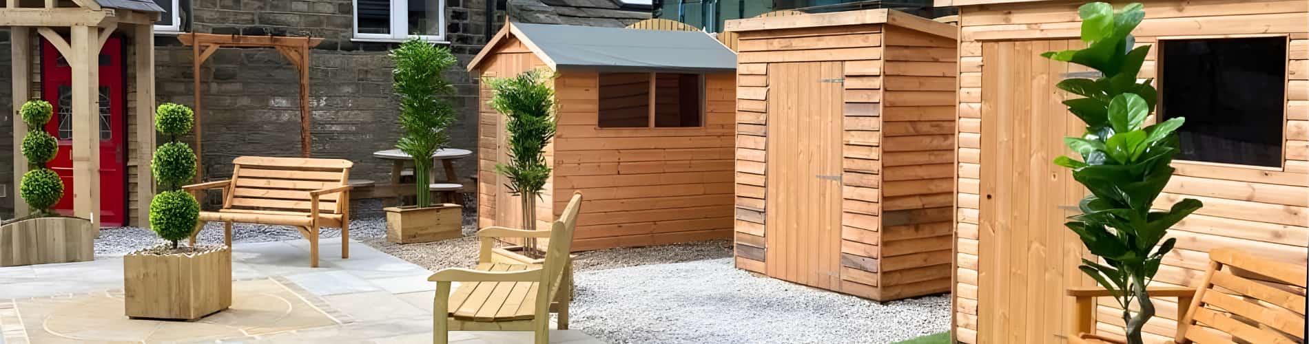 Outdoor display of wooden garden sheds, benches, and planters with neatly trimmed green topiary trees, set on gravel and stone paving, against a backdrop of a stone building.
