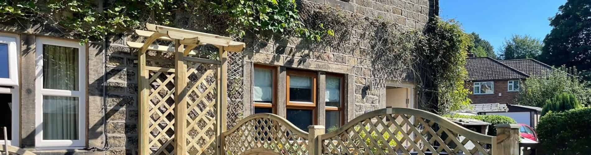 Stone house with white-framed windows, covered partially in ivy. In front, there is a wooden trellis fence and an arched trellis gate. Another house and trees are visible in the background under a clear blue sky.
