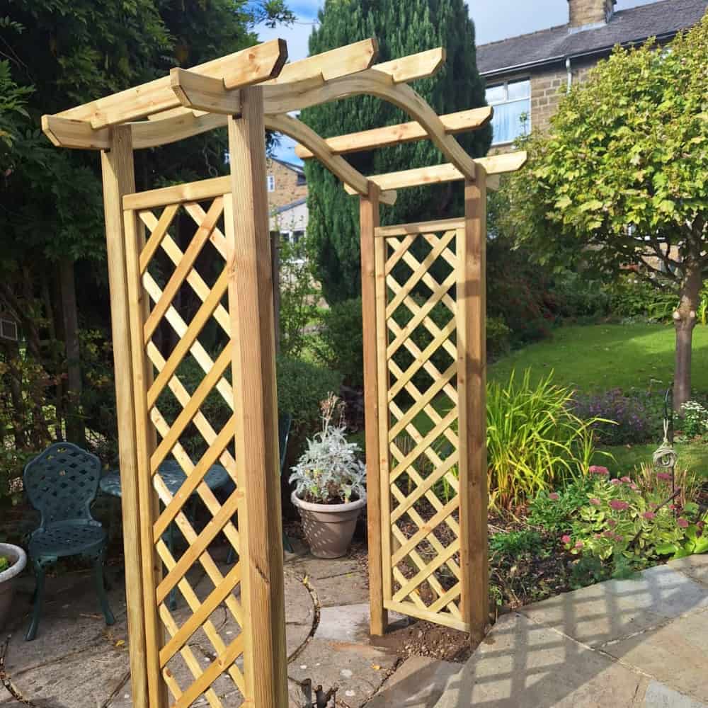 A wooden garden arbor with a lattice design stands on a stone patio, surrounded by green plants, trees, a potted plant, and outdoor furniture in a lush backyard garden.