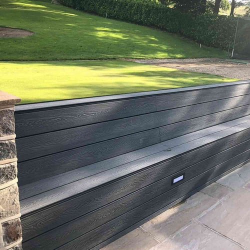 A stylish bench constructed from Bingley Fencing’s deX Classic Composite Decking Boards (3.6m, available in 4 colours) is integrated into a retaining wall next to a paved walkway, with grass and hedges behind. A small rectangular light embedded in the bench face adds a modern touch to this low-maintenance decking feature.