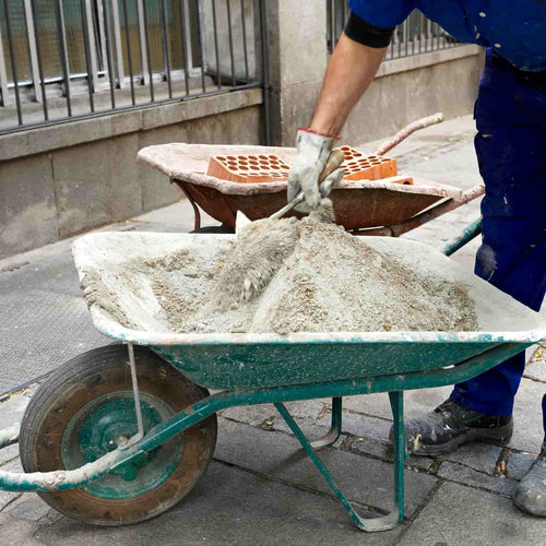 A construction worker in blue pants and gloves uses a shovel to mix Blue Circle Mastercrete Cement 25KG Bag - Plastic in a green wheelbarrow on the sidewalk; another wheelbarrow with bricks is seen behind.