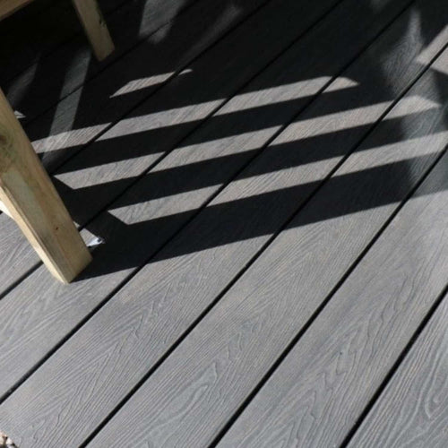 Bingley Fencing’s deX Classic Composite Decking Boards 3.6m, shown here in a stylish gray finish with realistic wood grain patterns, are partially shaded by diagonal striped shadows from a railing or fence. A wooden table or bench leg appears in the upper left corner of the image, highlighting the appeal of these low-maintenance decking boards for modern outdoor spaces.