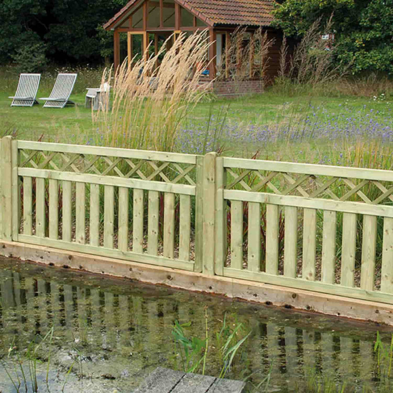 Load image into Gallery viewer, A KDM Cross Top Border Deck Panel (CT90) softwood fence with a lattice design stands beside a pond, complemented by tall ornamental grasses and a small lawn area with two lounge chairs in the background.
