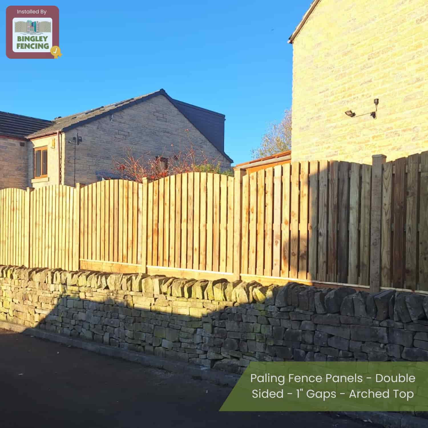 Bingley Fencingâ€™s Double Sided Paling Fence Panels with 1 gaps and arched tops are installed on a stone wall beside stone houses, casting shadows in bright sunlight. A Bingley Fencing sign appears at the top left.