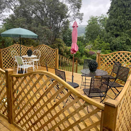 A wooden deck with lattice railing features two seating areas: one with a black metal table and chairs, and another with a white table and chairs under a green umbrella. A pink closed umbrella stands nearby. Trees surround the deck.
