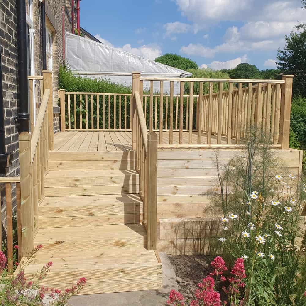 Wooden deck with stairs and railing next to a brick house, surrounded by a garden with flowers and greenery, under a blue sky with scattered clouds.