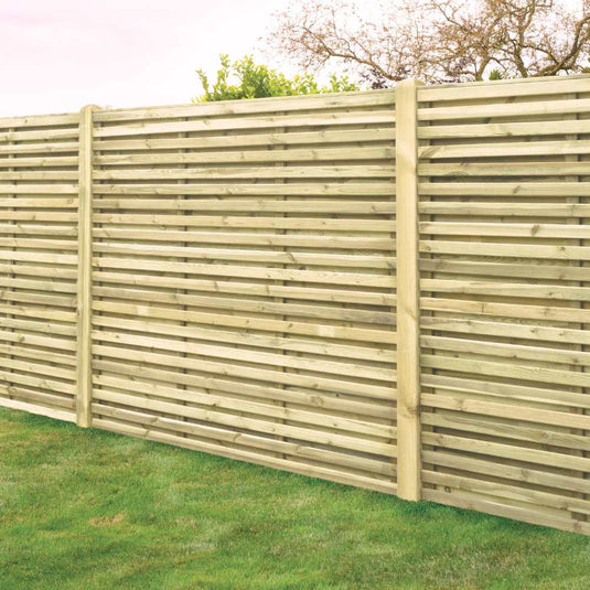 A KDM Double Sided Slatted European Fence (SDS180) stands on a lush, green lawn. Behind it, leafy trees are visible beneath a clear sky. The fence appears recently installed and shows no signs of wear.