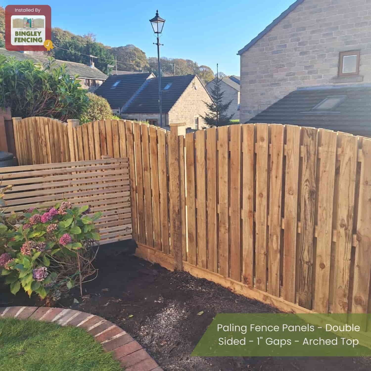 Bingley Fencingâ€™s Double Sided Paling Fence Panels with 1 gaps and arched tops are installed in a sunny garden with houses behind; a sign reads Installed by Bingley Fencing. Flowering plants grow nearby.