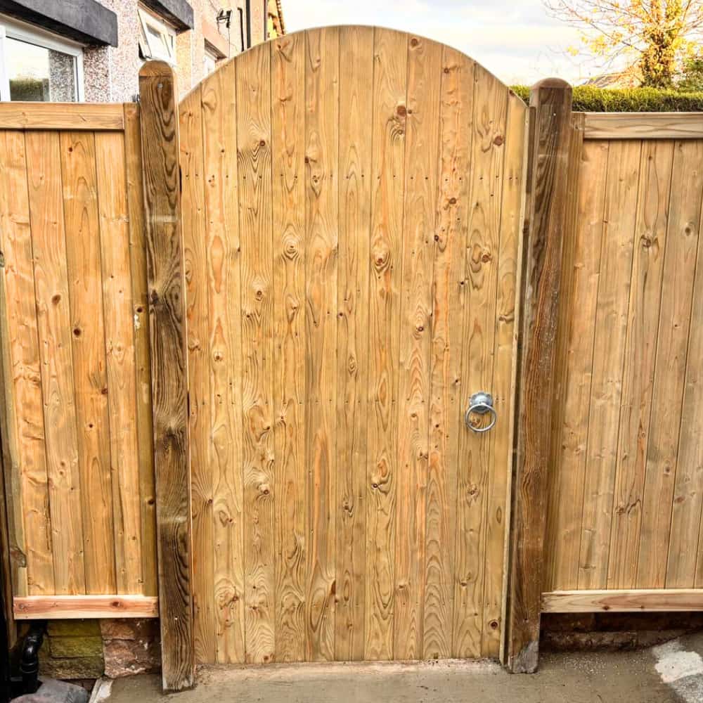 A wooden gate with an arched top is set between two vertical fence panels. The gate has visible wood grain, a silver handle on the right, and is framed by sturdy wooden posts.