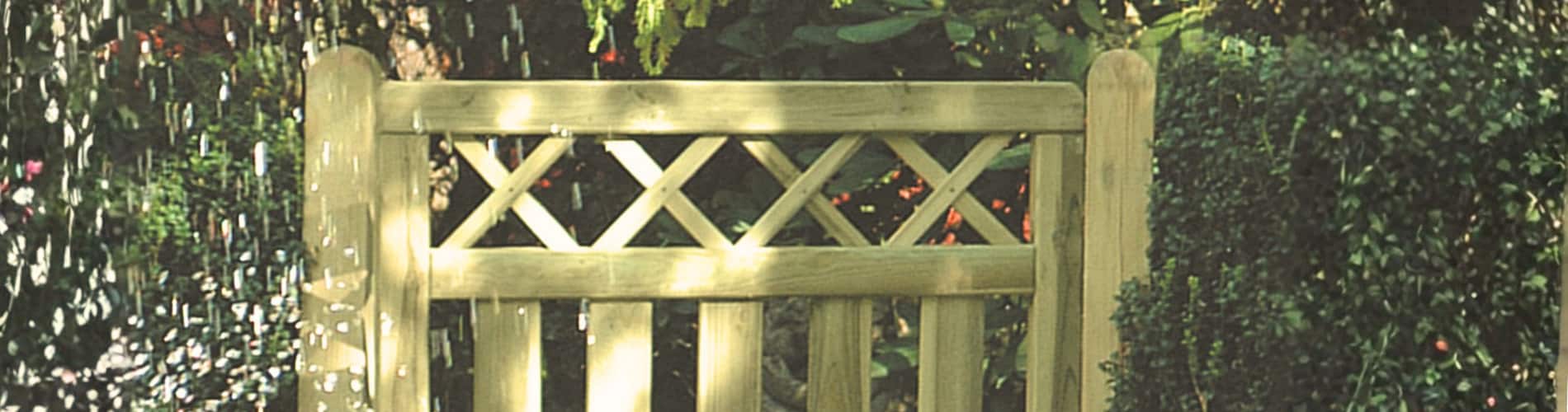A wooden garden gate with a crisscross pattern at the top, surrounded by green bushes and plants, with dappled sunlight filtering through.