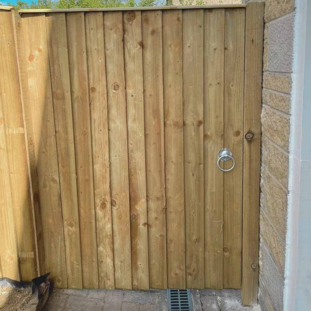 A wooden gate made of vertical planks with a round silver metal handle, set between a wooden fence and a stone wall, with a narrow drain on the ground in front.