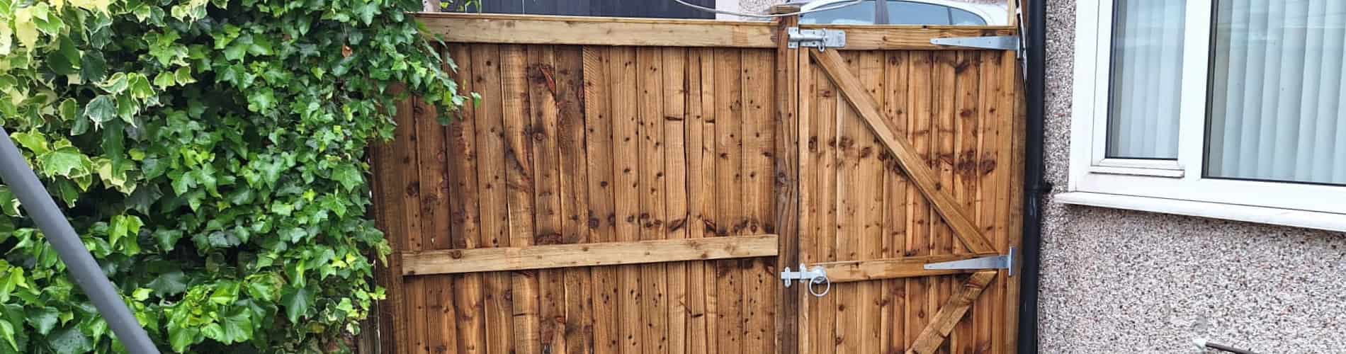 A wooden garden gate with metal hinges and a latch is installed between a wall covered in green ivy and the side of a pebble-dashed house with a window.