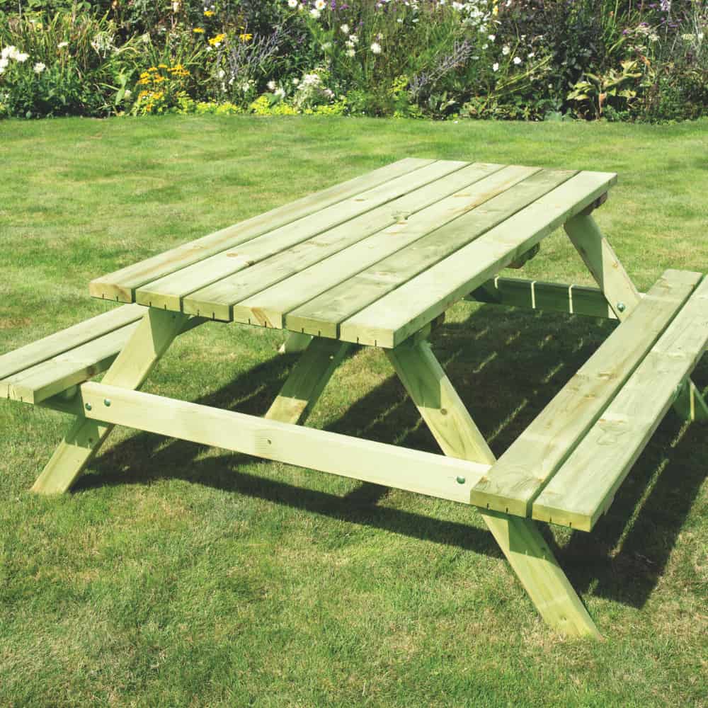 A light-colored wooden picnic table with attached benches sits on a grassy lawn, with a flowerbed and assorted plants in the background.