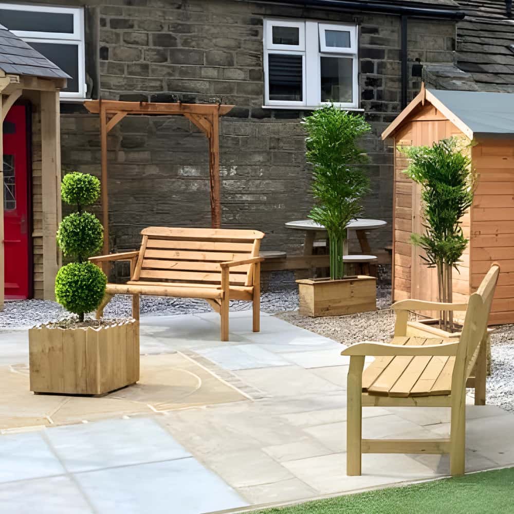 A tidy patio features wooden benches, a small wooden shed, potted topiary plants, and a paved stone floor, set against a stone building with white-framed windows.
