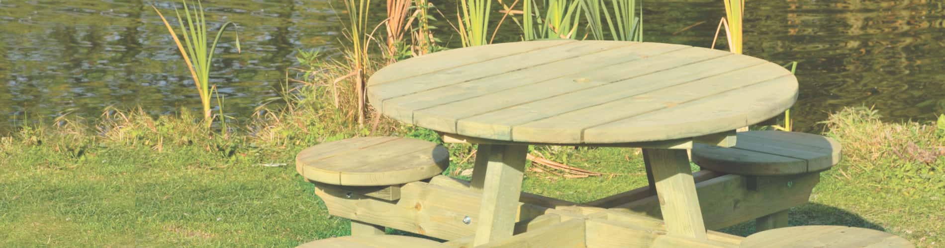 A wooden round picnic table with attached benches sits on grass near a pond with tall reeds and reflecting water.