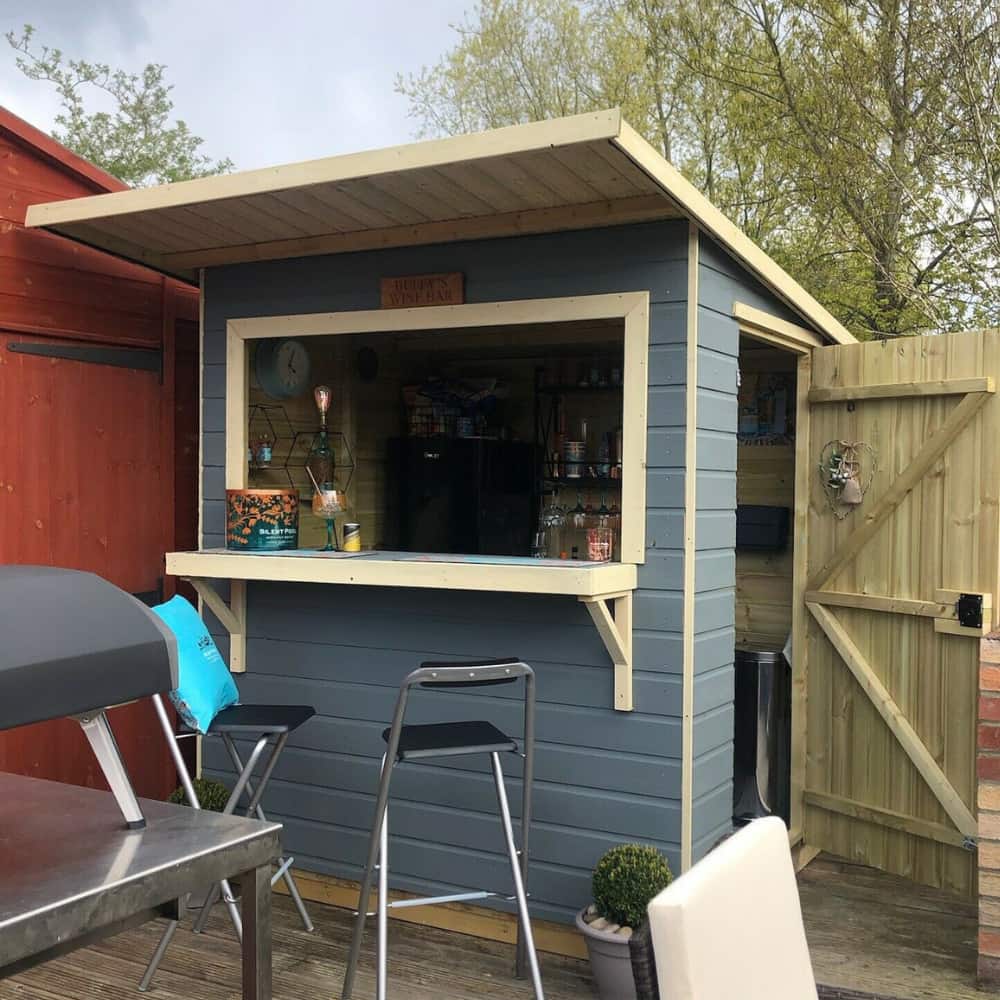 A small blue wooden garden bar shed with a counter, two stools, and an open serving window. The shed is next to a wooden fence and trees, with a BBQ grill in the foreground.