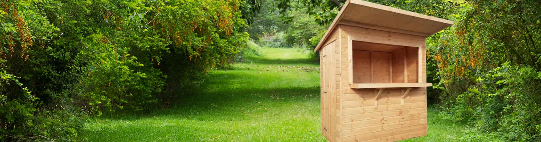 A small wooden kiosk with an open window stands on lush green grass, surrounded by dense trees and vibrant foliage in a forest clearing.