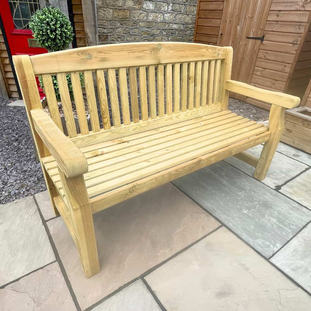 A light wood garden bench with slatted seat and backrest sits on a stone patio, next to a potted plant and a wooden shed.