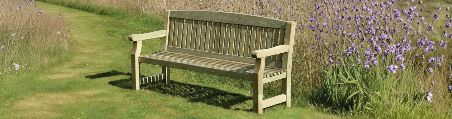 A wooden bench sits on a grassy path bordered by tall, wild grass and purple flowers under natural daylight.