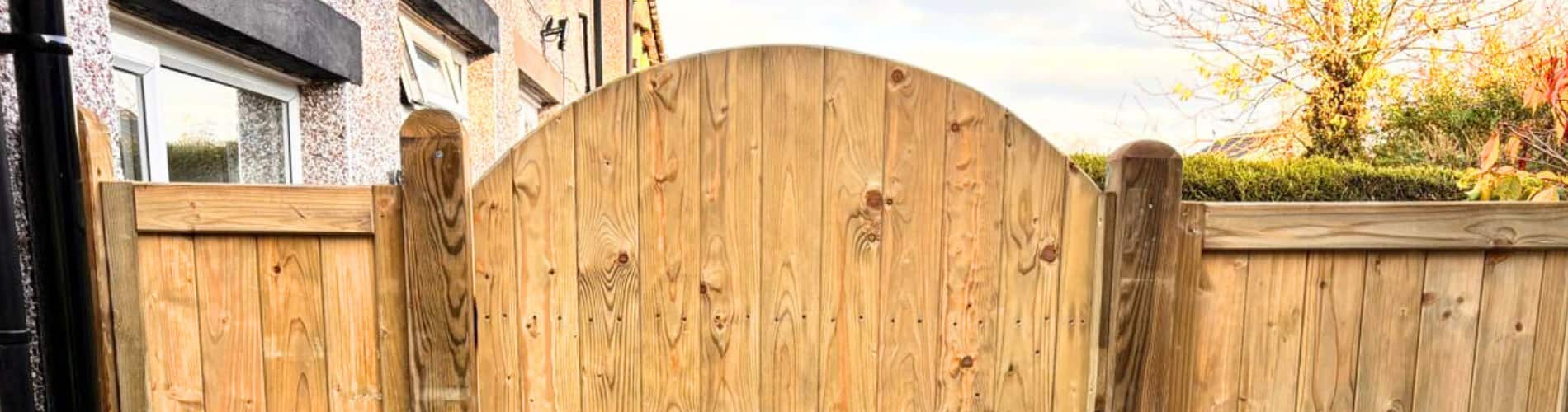 A wooden garden gate with an arched top is set between two matching fences in front of a house and trees under a partly cloudy sky.