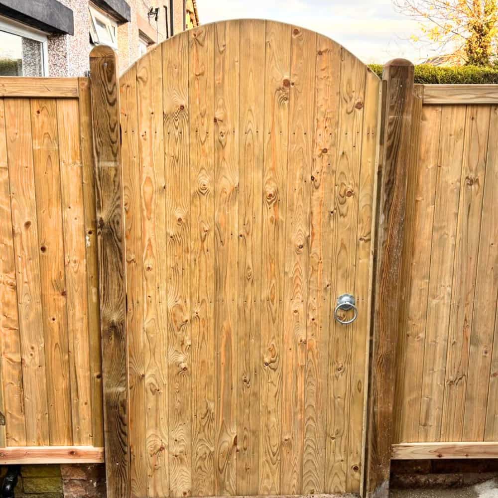 A wooden garden gate with a rounded top and vertical planks, flanked by a matching wooden fence. A metal handle is attached to the right side of the gate. Houses and trees are visible in the background.