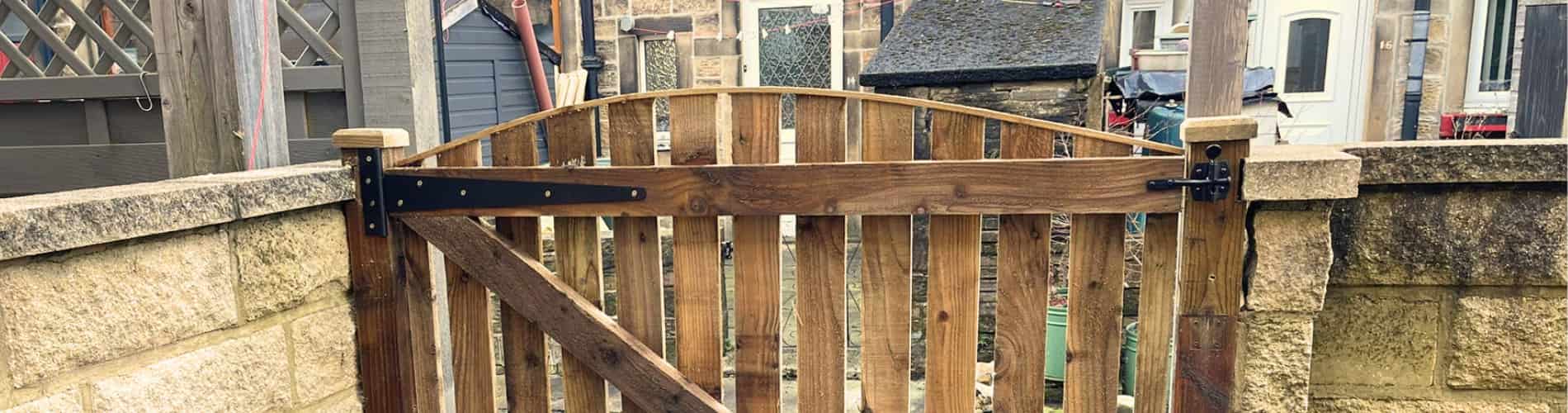 A wooden garden gate with vertical slats and black metal hinges is attached to stone walls, opening to a yard with sheds and houses in the background.