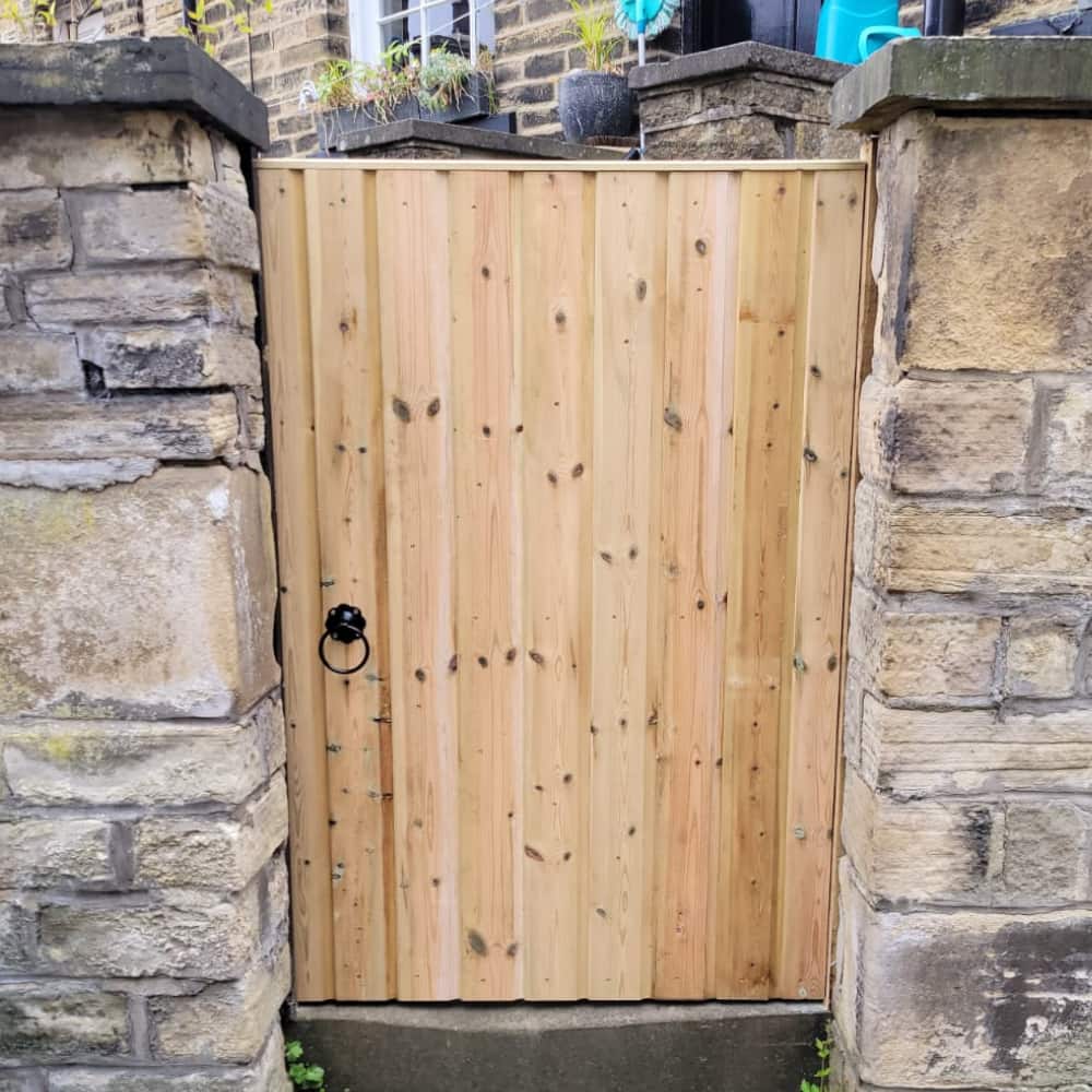 A wooden gate with vertical planks and a black metal ring handle is set between two stone walls outside a brick building with plants visible on top of the wall.
