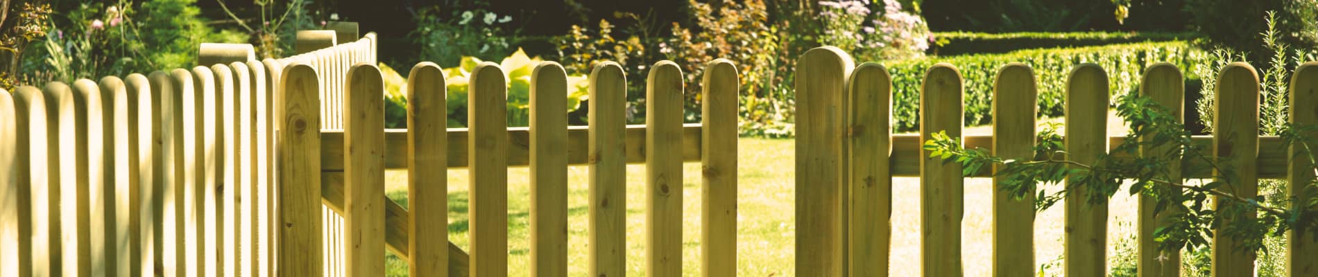 A wooden picket fence with an open gate stands in a sunlit garden, surrounded by green grass, bushes, and trees in the background.