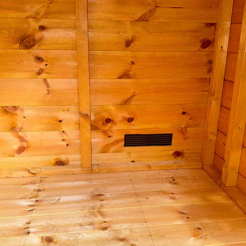 A close-up of a wooden cabin's varnished interior shows a Bingley Fencing Shed Air Vent (Louvre Vent with Fly Screen) installed on the lower wall, providing effective ventilation.