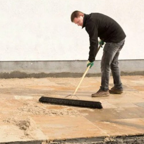A person in a black jacket and gloves spreads Joint-It Paving Jointing Compound 12.5kg by Joint It over Indian sandstone pavers on an outdoor patio using a large broom.