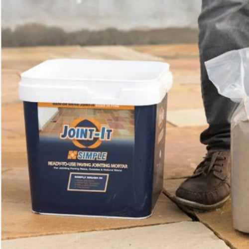 A blue and white tub labeled Joint It Paving Jointing Compound 12.5kg sits on stone paving beside a brown shoe and an open bag.