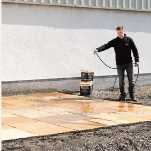 Someone uses a garden hose to water a newly laid patio after applying Joint It Paving Jointing Compound 12.5kg by Joint It. Two large containers are nearby on the unpaved ground, with a white wall in the background.