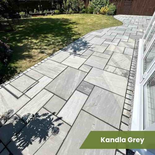 A modern patio features Bingley Fencing’s Indian Sandstone Paving Project Pack in mixed rectangles, bordered with bricks and leading to a shaded lawn. Sunlight filters through nearby plants, casting shadows on the natural stone surface.
