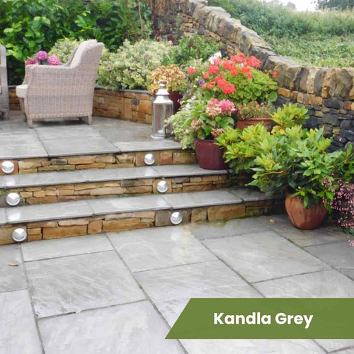 A natural stone patio with Bingley Fencing’s Indian Sandstone Paving Project Pack, potted plants, wicker furniture, and a stone wall with lit steps. A green banner in the bottom right reads “Kandla Grey.”.