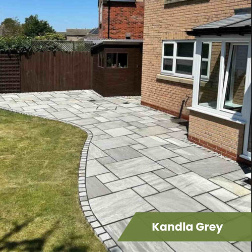 A backyard features a curved patio with Bingley Fencing's Indian Sandstone Paving Project Pack in Kandla Grey, adjacent to a lawn, brown wooden fence, small shed, and brick house. A green label in the corner says Kandla Grey.