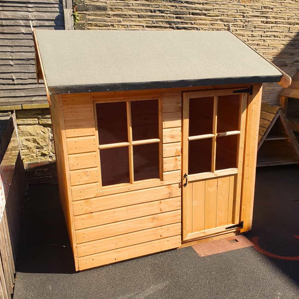 A small wooden playhouse with a slanted roof, two window panes, and a glass-paned door, placed on a paved surface next to brick and wooden fences.