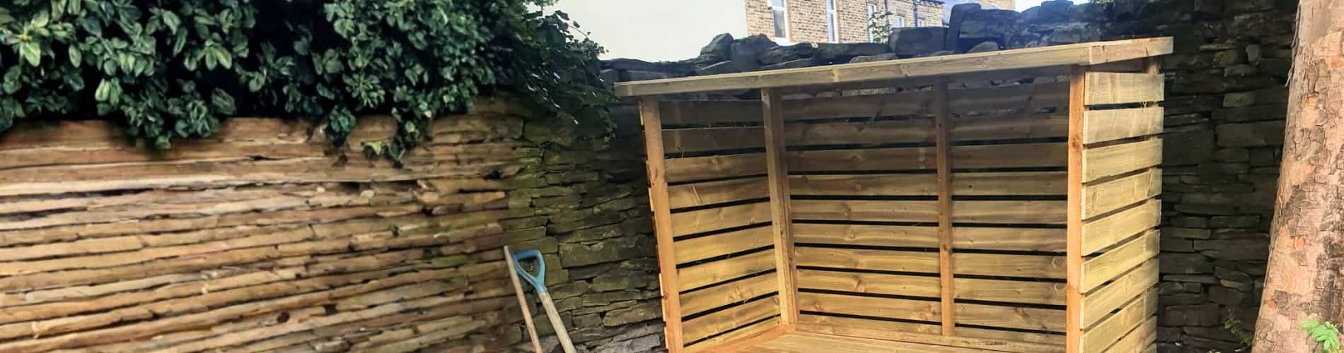 A wooden log store stands on a stone-paved area next to a stone wall, with a gardening spade leaning against the wall and greenery hanging over the corner.