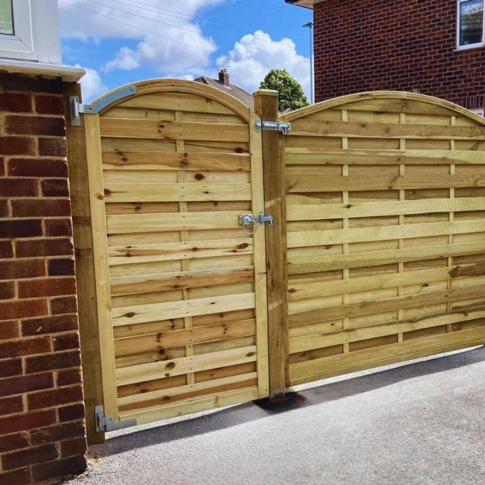 A wooden gate with an arched top is installed between two brick pillars, connected to a matching fence. The gate has horizontal wooden slats and metal hinges, with a house and blue sky in the background.