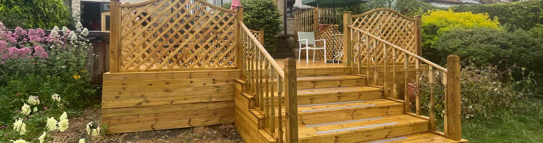 A wooden deck with stairs and handrails leads up to a garden area. The deck features lattice panels on either side, surrounded by plants, flowers, and greenery in a backyard setting.