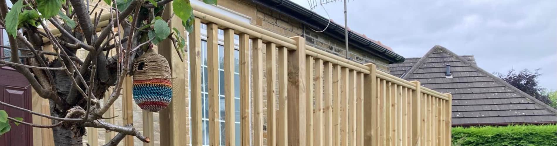 A wooden fence with vertical slats stands beside a tree with a multicolored yarn-wrapped hanging ornament, next to a stone house with a tiled roof and cloudy sky in the background.