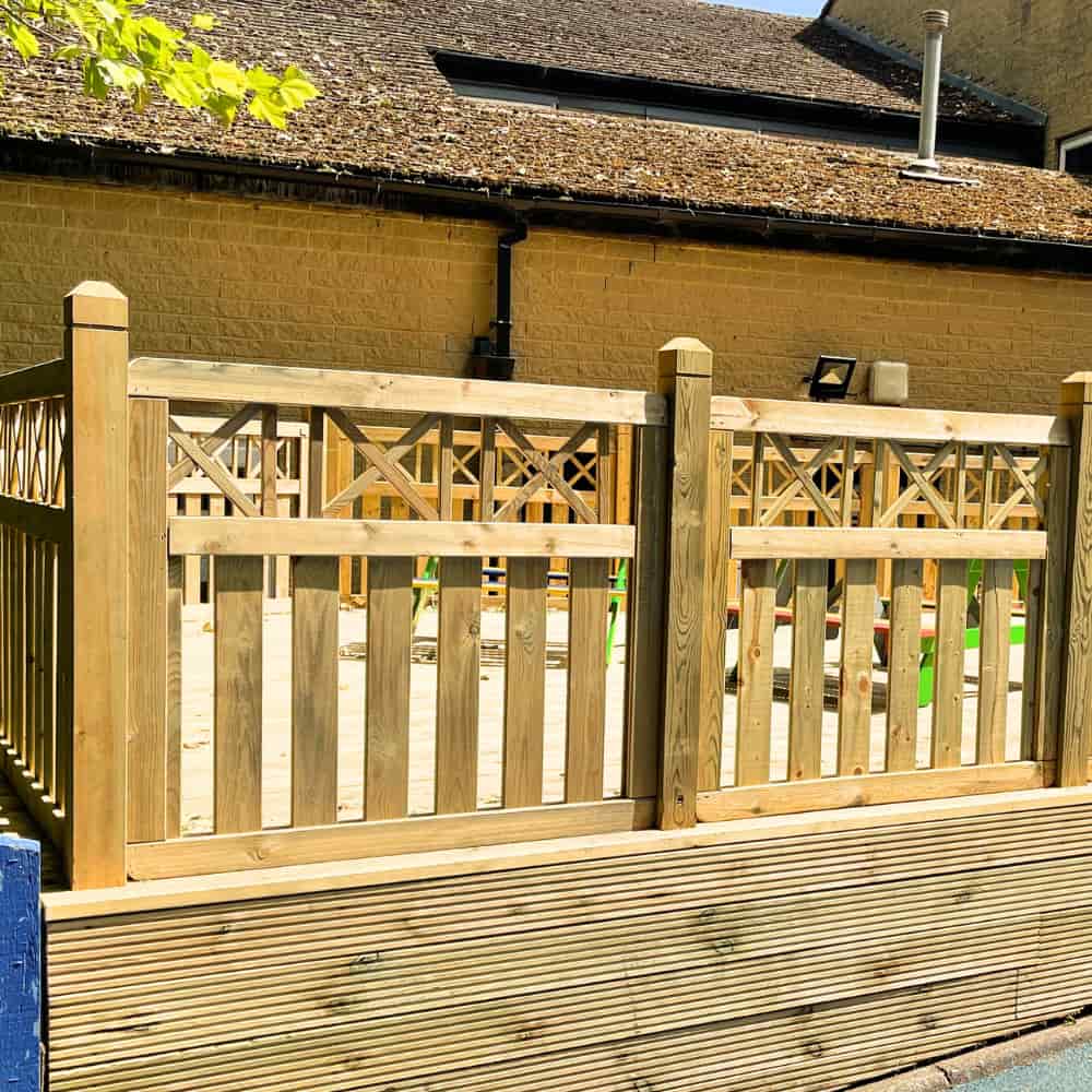A wooden fence with decorative cross patterns encloses a sandy play area, set against a yellow brick building with a sloped roof on a sunny day.