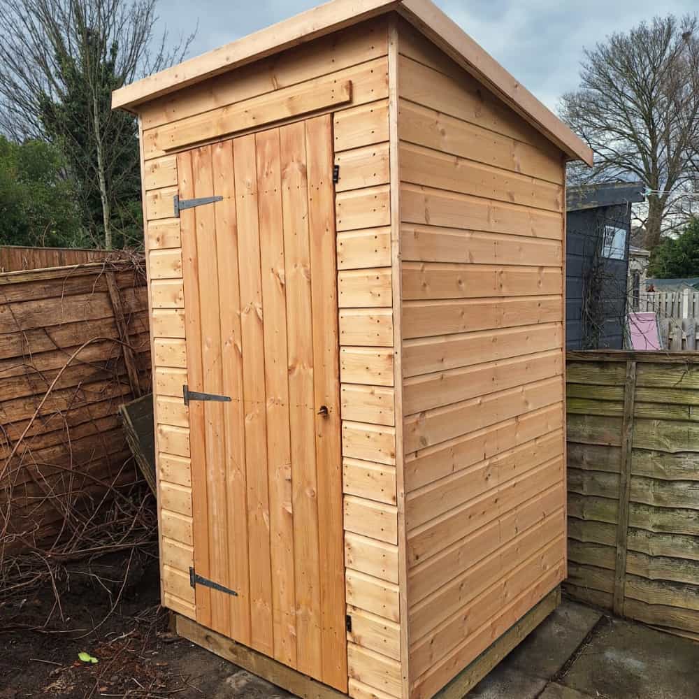 A new wooden garden shed with a slanted roof and metal hinges stands on a paved area next to a wooden fence in an outdoor garden setting.