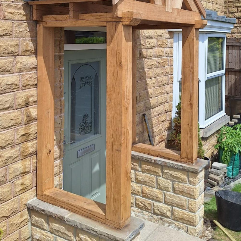 Load image into Gallery viewer, A light green door with decorative glass panels sits in a stone house beneath the Traditional Oak Porch &amp; Frame by Bingley Fencing, crafted with classic joinery. Sunlight casts shadows on the brickwork, and theres a window and plants by the wall.
