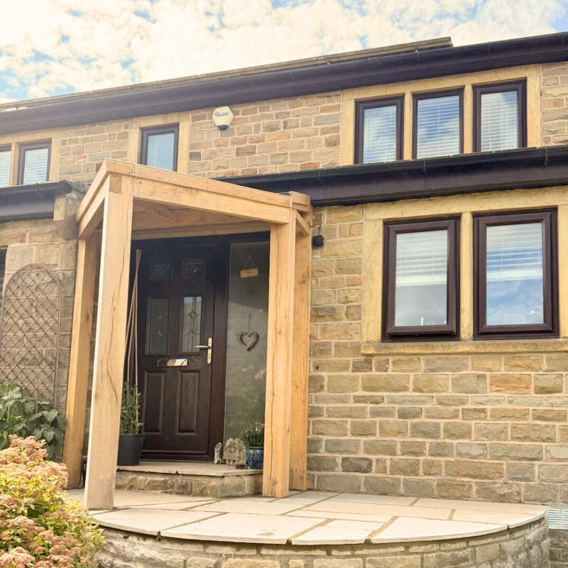 Load image into Gallery viewer, A stone house with a dark wooden front door and rectangular windows with white blinds features the Bingley Fencing Oak Entrance Porch and potted plants at the entrance. The sky above is partly cloudy.
