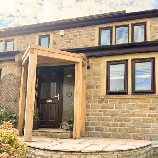 A stone house with a dark wooden front door and rectangular windows with white blinds features the Bingley Fencing Oak Entrance Porch and potted plants at the entrance. The sky above is partly cloudy.