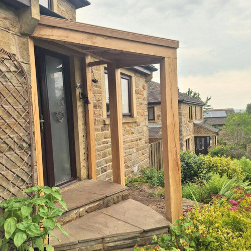 A stone house features a Bingley Fencing Oak Entrance Porch framing the entry, surrounded by lush greenery and a tidy garden, with another house in the background beneath a cloudy sky.