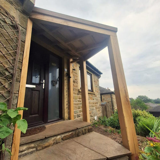 A stone house entrance features Bingley Fencing’s Oak Entrance Porch, supported by wooden beams and a dark wood door, surrounded by greenery and plants beneath an overcast sky.
