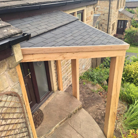 The Bingley Fencing Oak Entrance Porch features a sloped shingle roof and sturdy wooden posts, offering covered protection for your stone pathway and entrance door, beautifully complementing your home’s exterior.