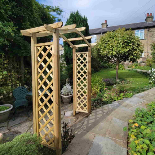A KDM Omega Top Arch, made from tanalised softwood with an intricate arch design, stands elegantly on a pathway surrounded by lush greenery and tall grasses. This stunning garden feature is beautifully set against a backdrop of trees and fields under a partly cloudy sky.